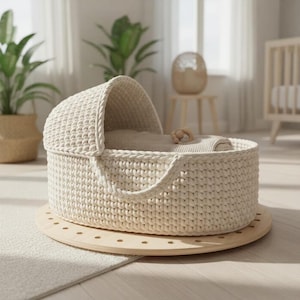 Könnte beinhalten: Eine cremefarbene, handgehäkelte Babywiege mit einer abgerundeten Haube und einem Griff. Die Wiege steht auf einer kreisförmigen Holzbasis. Im Inneren sind eine kleine Decke und ein Holzspielzeug zu sehen. Die Wiege befindet sich in einem hellen, luftigen Raum.