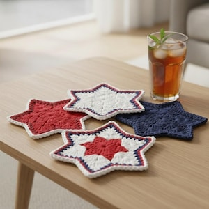 May include: Four star-shaped crochet coasters in red, white, and navy blue sit on a wooden table. One coaster has a red center, white middle, and navy blue border. A glass of iced tea with a mint sprig is also on the table.