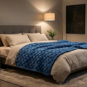 May include: A bed featuring a thick, chunky knit blue throw blanket draped over beige bedding. The bed has a gray tufted headboard and is positioned next to a wooden nightstand with a small lamp. The room includes a floor lamp and wall sconce.