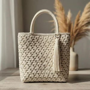 May include: Cream-colored, hand-crocheted tote bag with a textured stitch pattern. The bag features two rounded handles and a decorative tassel. The bag is sitting on a wooden surface.