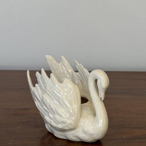 May include: A white ceramic swan figurine with detailed wing and feather textures. The swan has a curved neck and a small opening on its back, suggesting it may be a decorative planter or holder.