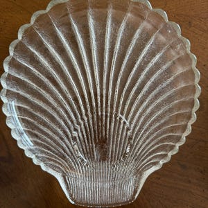May include: A clear glass seashell-shaped dish with a scalloped edge. The dish has a ribbed texture, with lines radiating from the center to the edge, resembling the natural pattern of a seashell. The dish is designed for decorative or serving purposes.