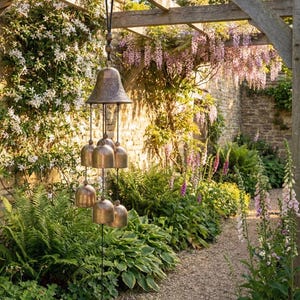 Könnte beinhalten: Ein bronzefarbener Windspiel mit einer großen Glocke oben und kleineren Glocken, die darunter hängen. Das Windspiel ist an einer Holzpergola aufgehängt, umgeben von üppigem Grün und blühenden Pflanzen. Am Boden hängt eine dekorative Münze.