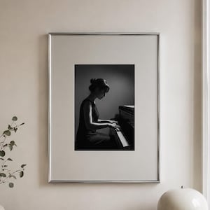 Könnte beinhalten: Ein gerahmtes Schwarz-Weiß-Foto, das eine Person beim Klavierspielen zeigt, präsentiert in einem dünnen silbernen Rahmen. Das Kunstwerk hängt an einer hellen Wand über einem Holzkonsolentisch mit einer Keramikvase, einer Schale und einem Buch.