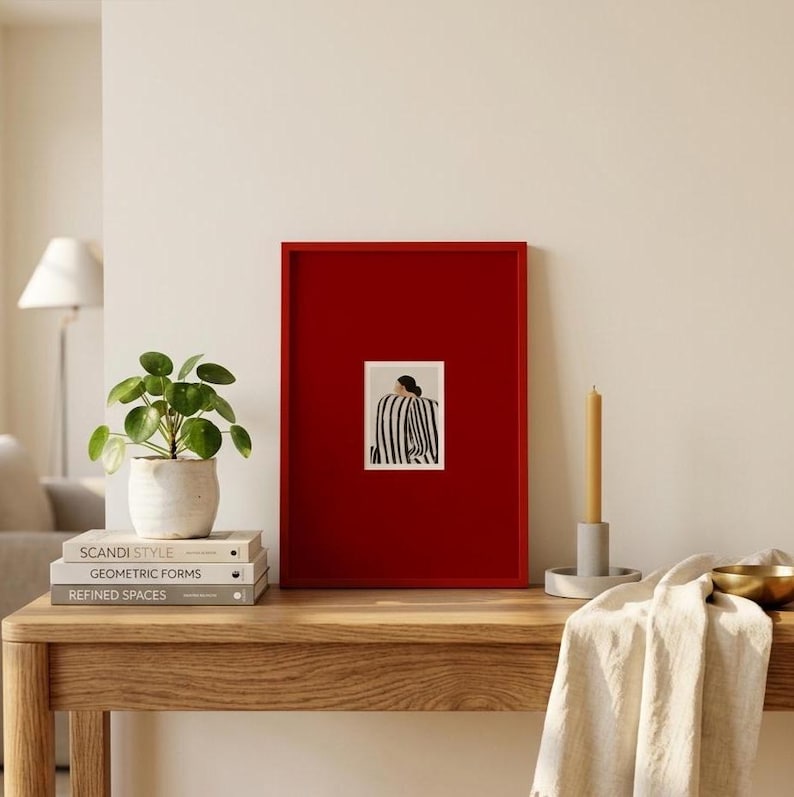 Op de afbeelding: Een rode fotolijst met een kunstafdruk met een zwart-wit gestreepte figuur. De afdruk staat op een houten tafel met een kleine potplant, boeken, een kaars en een gouden kom. De boeken heten "Scandi Style", "Geometric Forms" en "Refined Spaces".