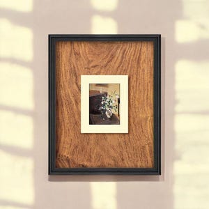 May include: A framed artwork featuring a still life painting of flowers in a vase. The painting is set within a cream-colored mat and a dark wooden frame. The frame is set against a textured, brown wooden background.