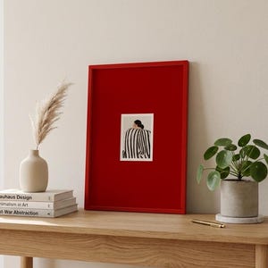 Op de afbeelding: Een rood ingelijste kunstprint met een zwart-wit gestreepte figuur. De kunst is te zien op een houten tafel, met een vaas met gedroogd pampasgras, boeken en een potplant. Minimalistisch interieur.