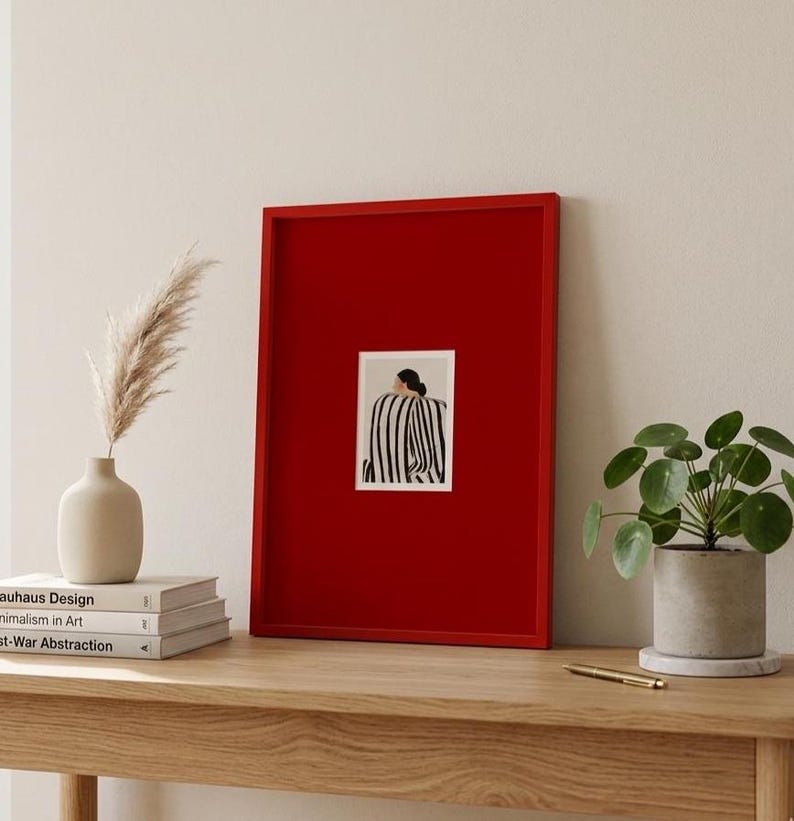 Op de afbeelding: Een rood ingelijste kunstprint met een zwart-wit gestreepte figuur. De kunst is te zien op een houten tafel, met een vaas met gedroogd pampasgras, boeken en een potplant. Minimalistisch interieur.