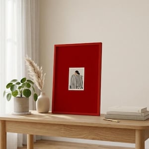 Op de afbeelding: Een ingelijst kunstwerk met een rode rand. Het kunstwerk heeft een kleine rechthoekige print met een zwart-wit gestreept ontwerp. Het frame wordt tentoongesteld op een houten tafel, samen met een potplant en een decoratieve vaas.