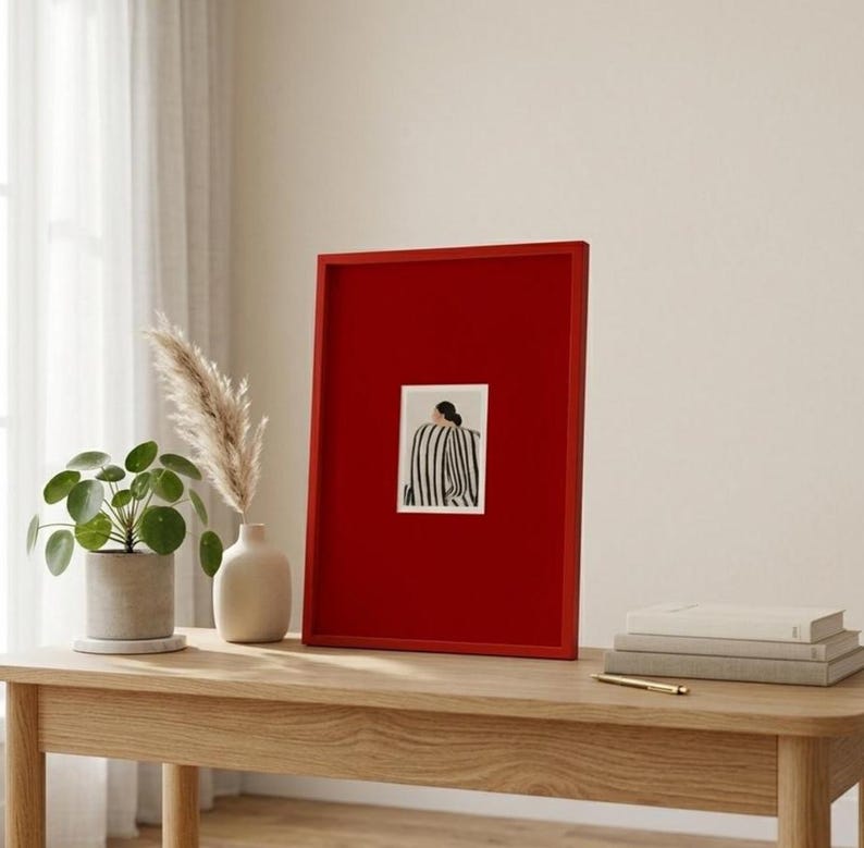 Op de afbeelding: Een ingelijst kunstwerk met een rode rand. Het kunstwerk heeft een kleine rechthoekige print met een zwart-wit gestreept ontwerp. Het frame wordt tentoongesteld op een houten tafel, samen met een potplant en een decoratieve vaas.