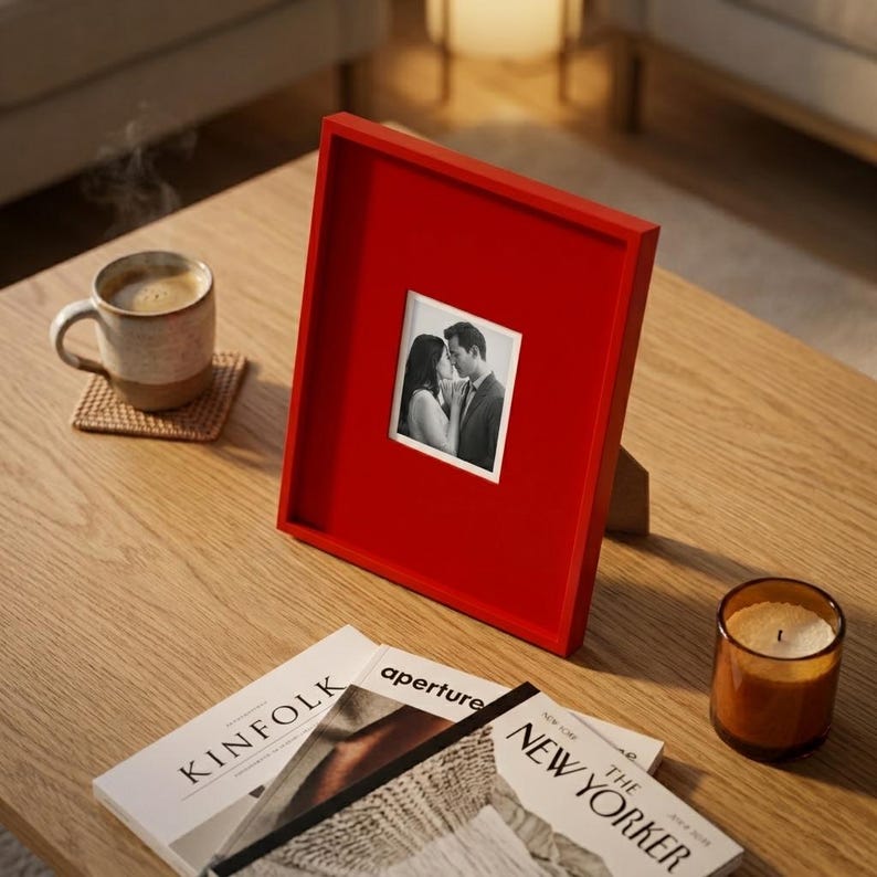 Op de afbeelding: Een rode vierkante fotolijst met een zwart-witfoto van een stel. Een koffiemok en een kaars staan op de houten tafel, samen met tijdschriften.