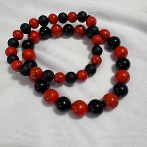 May include: Two beaded bracelets. One bracelet features red and black beads, while the other has red and black beads. The beads are round and appear to be made of stone. The bracelets are displayed on a white surface.