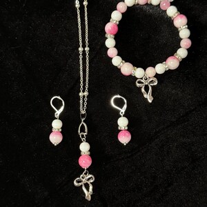 May include: A jewelry set featuring a necklace, bracelet, and earrings. The necklace and earrings have pink and white beaded accents, with a silver-tone charm. The bracelet has similar beads and a silver-tone bow charm. The jewelry is displayed on a black surface.