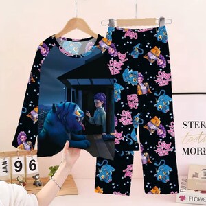 May include: Black pajama set with a long-sleeved top and pants. The top features a graphic of a blue tiger and a person. The pants have a repeating pattern of the tiger, a person, and pink text. The sleeves have a repeating pattern of the tiger and a person.