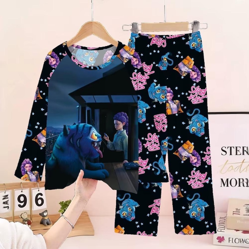 May include: Black pajama set with a long-sleeved top and pants. The top features a graphic of a blue tiger and a person. The pants have a repeating pattern of the tiger, a person, and pink text. The sleeves have a repeating pattern of the tiger and a person.