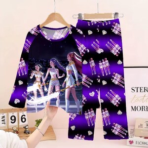 May include: A two-piece pajama set featuring a long-sleeved top and pants. The top has a dark purple background with a graphic of three figures. The pants have a repeating pattern of figures and diamonds on a purple and black background.