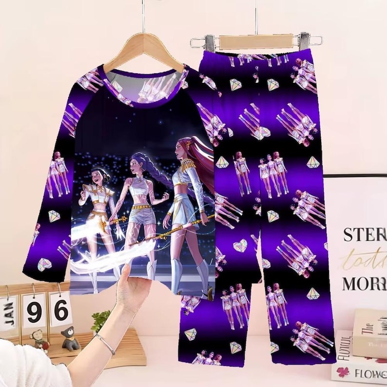 May include: A two-piece pajama set featuring a long-sleeved top and pants. The top has a dark purple background with a graphic of three figures. The pants have a repeating pattern of figures and diamonds on a purple and black background.