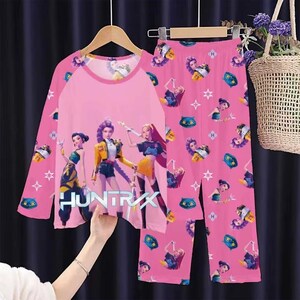 May include: Pink pajama set featuring a long-sleeved top and matching pants. The top has a pink body with a graphic of three animated figures and the word "HUNTRIXX". The pants have a repeating pattern of animated figures, stars, and other graphics.