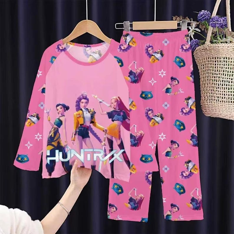 May include: Pink pajama set featuring a long-sleeved top and matching pants. The top has a pink body with a graphic of three animated figures and the word "HUNTRIXX". The pants have a repeating pattern of animated figures, stars, and other graphics.