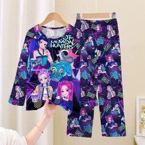 May include: A long-sleeved pajama set featuring a vibrant graphic design. The top and pants are covered in illustrations of anime-style characters, a blue tiger, and the text "OF DEMON HUNTERS" in white. The set is primarily purple with neon accents.