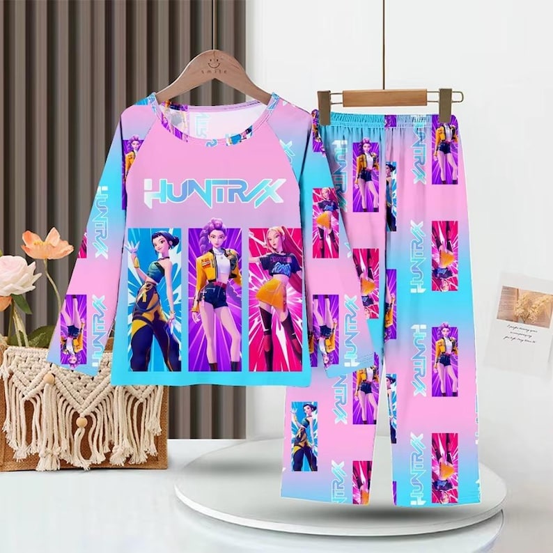 May include: A two-piece pajama set featuring a long-sleeved top and pants. The set is predominantly pink and blue with a repeating graphic of stylized figures. The top displays the word "HUNTRAX".