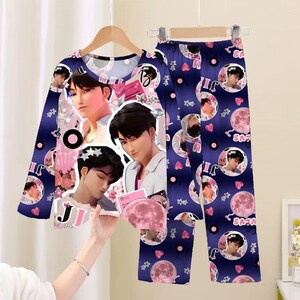 May include: Navy blue pajama set featuring a long-sleeved top and pants. The set is adorned with multiple images of a person, pink moons, hearts, and stars. The top has a round neckline and the pants have an elastic waistband.