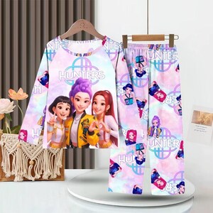 May include: A two-piece pajama set featuring a long-sleeved top and pants. The top displays three cartoon characters with the word "Hunters". The pants have a repeating pattern of the same characters. The pajamas are in shades of pink, purple, and blue.