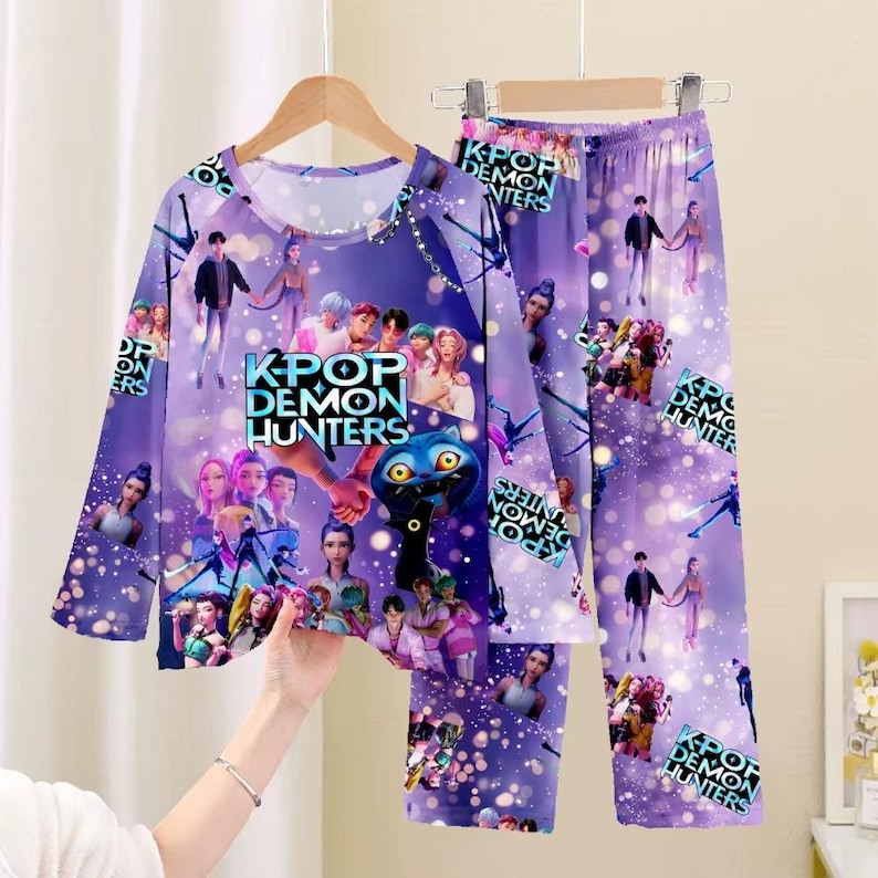May include: A purple pajama set with a long-sleeved top and pants. The set features a repeating pattern of figures and the text "KPOP DEMON HUNTERS". The pajama set is hanging on a wooden hanger.