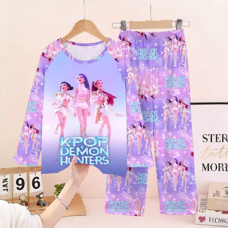 May include: A two-piece pajama set featuring a long-sleeved top and pants. The top is lavender with a graphic of three figures and the text "KPOP DEMON HUNTERS." The pants have a repeating pattern of the same figures. The pajamas are hanging on a wooden hanger.