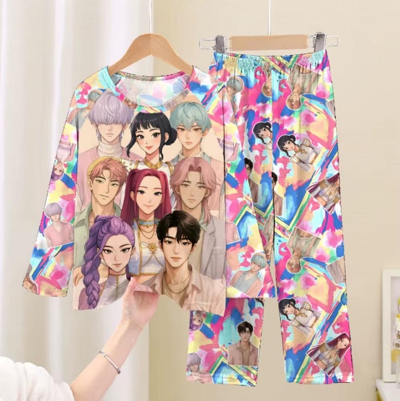 May include: A colorful pajama set featuring a long-sleeved top and matching pants. The set is adorned with a vibrant print of anime-style characters. The top is hanging on a wooden hanger. The pants have an elastic waistband.