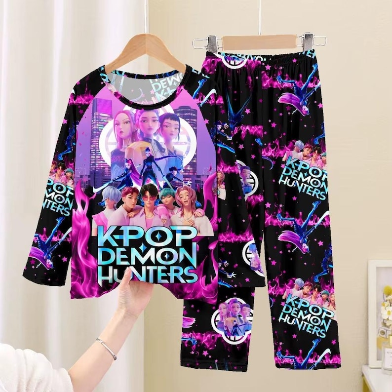 May include: A long-sleeved pajama set featuring a vibrant graphic. The top displays a group of people with the text "KPOP DEMON HUNTERS" over a purple and pink flame design. The matching pants have a repeating pattern with the same text.