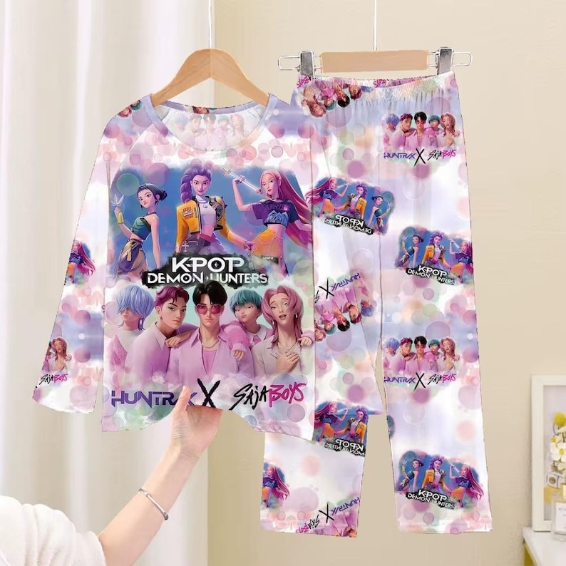 May include: A pajama set featuring a long-sleeved top and pants. The set is white with a colorful graphic of a K-pop group and the text "KPOP DEMON HUNTERS", "HUNTR X SAJA BOYS". The design includes pink and purple accents.