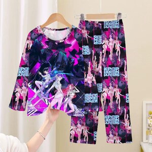 May include: A long-sleeved pajama top and matching pants set. The set features a vibrant graphic design with anime-style characters in shades of pink, purple, and black. The text "K-POP DEMON HUNTERS" is visible on the clothing.