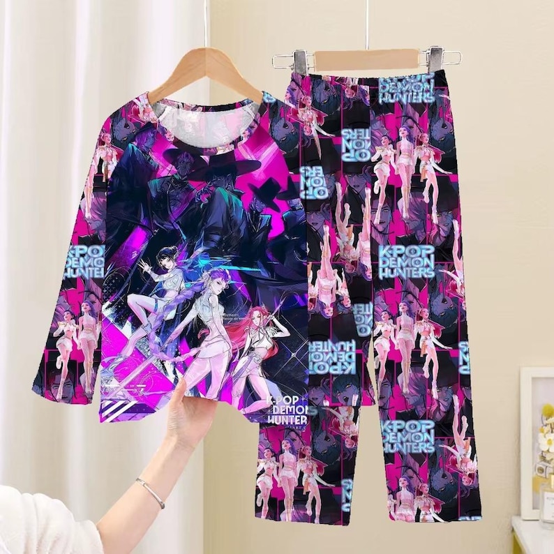 May include: A long-sleeved pajama top and matching pants set. The set features a vibrant graphic design with anime-style characters in shades of pink, purple, and black. The text "K-POP DEMON HUNTERS" is visible on the clothing.