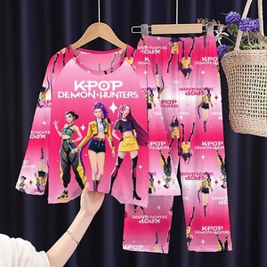 May include: Pink K-Pop Demon Hunters pajama set. The long-sleeved top and pants feature a vibrant graphic of three anime-style characters and the text "K-Pop Demon Hunters". The pajamas are hanging on a wooden hanger.