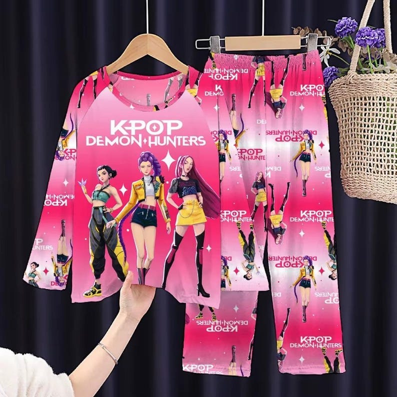 May include: Pink K-Pop Demon Hunters pajama set. The long-sleeved top and pants feature a vibrant graphic of three anime-style characters and the text "K-Pop Demon Hunters". The pajamas are hanging on a wooden hanger.
