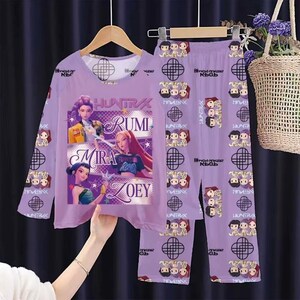 May include: A lavender pajama set featuring a long-sleeved top and matching pants. The top displays the text "HUNTRA" and the names "RUMI", "MIRA", and "ZOEY" with images of animated characters. The pants have a repeating pattern of cartoon figures and geometric shapes.
