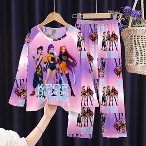 May include: A two-piece pajama set featuring a long-sleeved top and pants. The set is predominantly pink and purple, with a repeating pattern of anime-style characters. The top displays three characters and the text "KPOP DEMON HUNTERS".