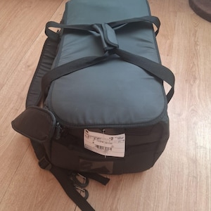 May include: A black and gray padded bag with a top handle and shoulder straps. The bag has a rectangular shape with a zippered compartment and a label attached. The bag is designed for carrying items.