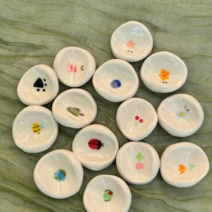以下が含まれることがあります： 小さな白い陶器の皿のコレクション。各皿には、花、昆虫、さまざまな色の抽象的な形など、ユニークな手描きのデザインが施されています。皿は緑色の布の表面に配置されています。