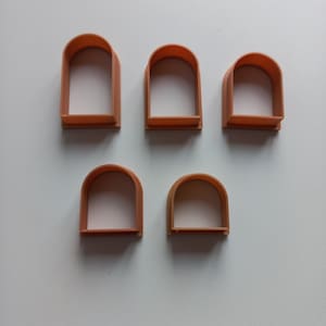 Pode incluir: Cinco cortadores de biscoitos em forma de arco de cor laranja. Os cortadores estão dispostos em um grupo de cinco, com dois na parte inferior e três na parte superior. Os cortadores são feitos de plástico e têm um acabamento liso.