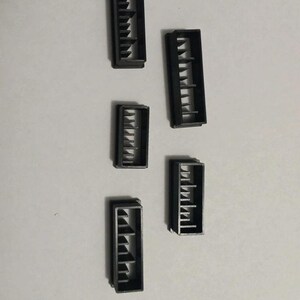 Puede incluir: Vista cenital de cinco pequeños componentes rectangulares de plástico negro. Cada componente tiene una serie de ranuras verticales paralelas. Los componentes están dispuestos sobre una superficie gris claro.