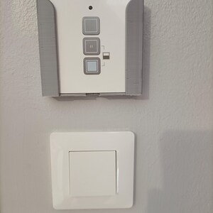 May include: A white control panel with three gray buttons and a small indicator light, mounted on a gray bracket. Below, a white light switch with a square button is installed on a light gray wall.