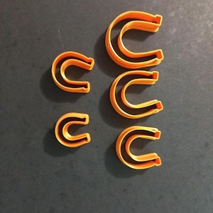 Puede incluir: Cinco cortadores de galletas de plástico naranja con forma de letra C de varios tamaños están dispuestos sobre una superficie gris oscuro. Los cortadores tienen un interior hueco y una forma redondeada, para hornear o manualidades.