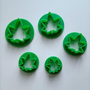 Puede incluir: Cinco cortadores de galletas verdes con forma de hoja de diferentes tamaños. Cada cortador presenta un diseño de hoja detallado dentro de un marco circular. Los cortadores están hechos de un material verde sólido y están dispuestos sobre una superficie blanca.