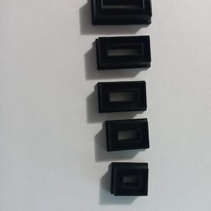 May include: Five black rectangular plastic components with a recessed center, arranged vertically on a white surface. Each component has a shadow, suggesting a three-dimensional design. The objects appear to be small, possibly parts of a larger assembly.