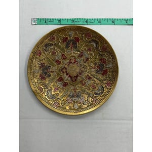 May include: A round, gold-colored decorative plate with an intricate floral design in red, black, and silver. The plate is approximately 6 inches in diameter. The plate has a detailed, embossed pattern around the edge.