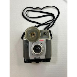 May include: A vintage Kodak Brownie Flashmite 20 camera with a black strap. The camera has a silver face with a lens and a flashbulb. The camera has the Kodak logo and the words "Brownie Flashmite 20" printed on the front.