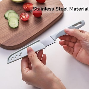 May include: A stainless steel knife with a light blue handle and sheath. The knife is being held by a person. The text "Stainless Steel Material" is visible in the image. Sliced cucumbers and tomatoes are on a wooden cutting board.