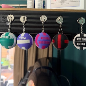 May include: Five spherical netball keychains hanging from a black shelf. The keychains feature various colours including teal, purple, pink, black, and white, with text printed on each: NETBALLER, STORM, NETBALL OBSESSED, and NETBALL QUEEN.